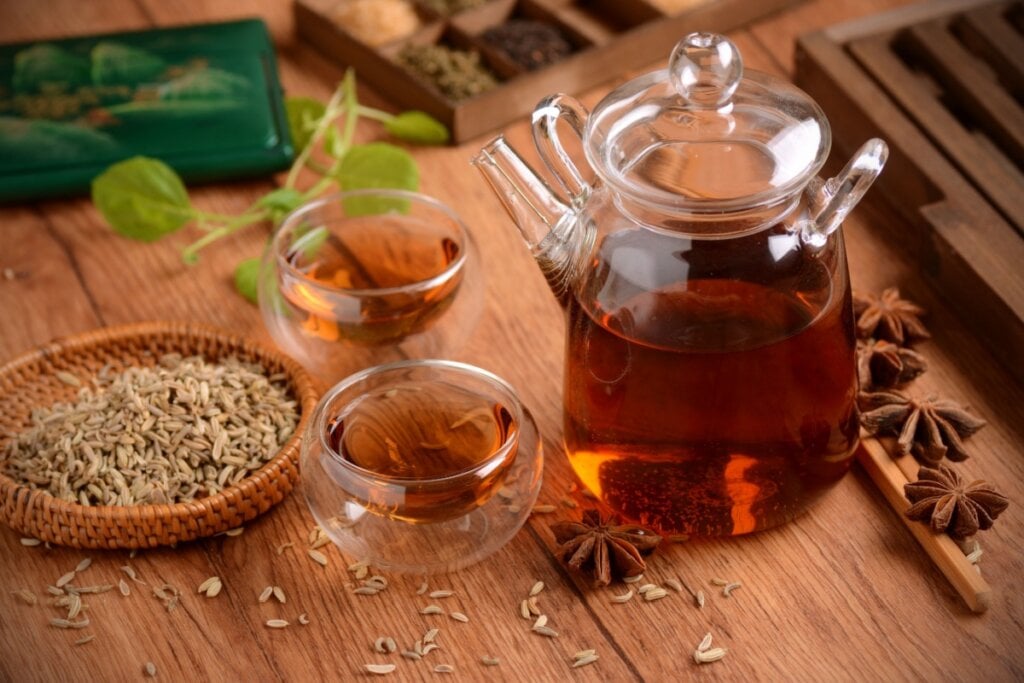 Chá de erva-doce com anis-estrelado em bule de vidro e servido em dois copos de vidro em cima de mesa de madeira, com sementes de erva-doce e anis-estrelado espalhados na mesa